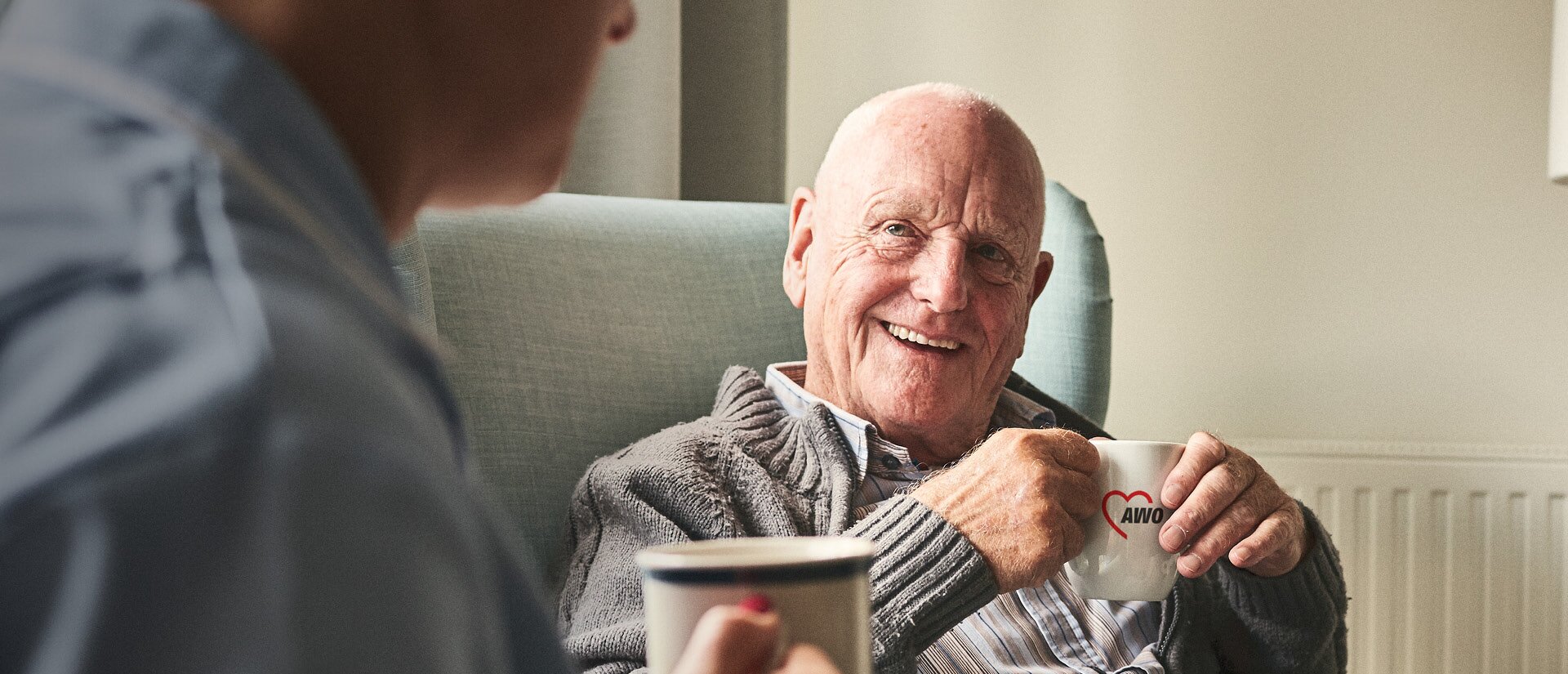 Das Foto zeigt einen alten Mann, der entspannt und glücklich in einem Sessel sitzt. In seiner Hand hält er eine AWO-Tasse.
